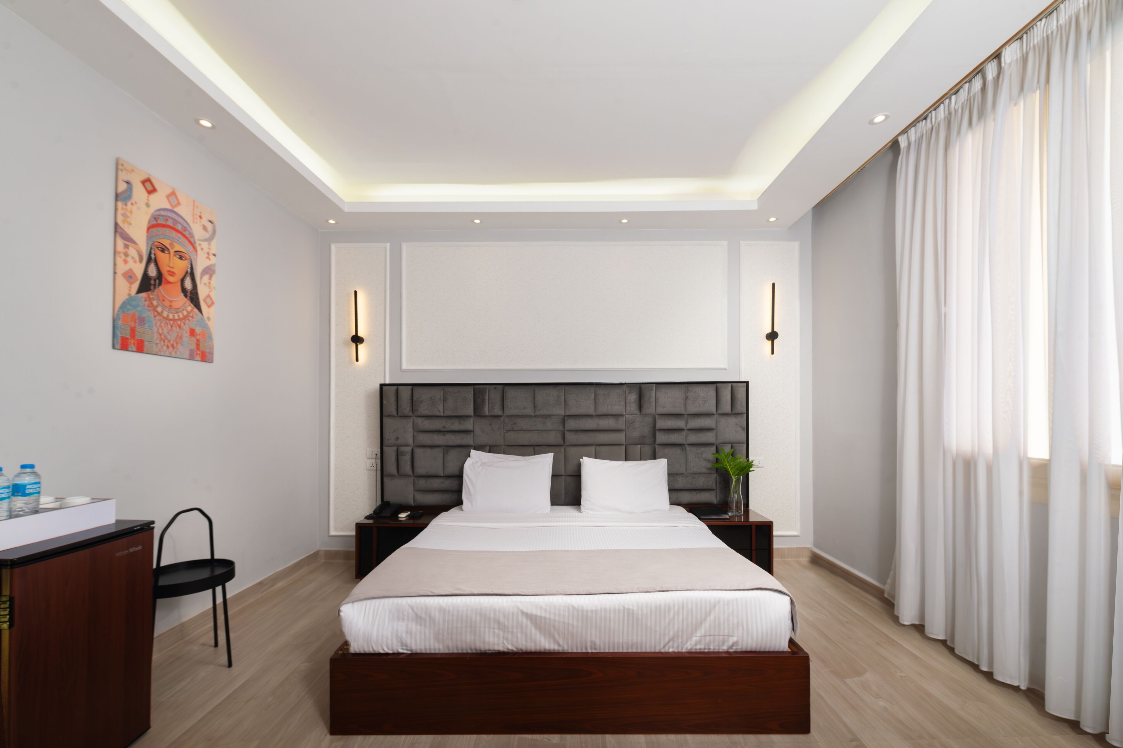 A modern hotel room with a large bed, a desk with a chair, a nightstand, and a wall-mounted artwork.