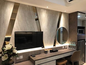 A modern living room with a large flat-screen TV, a round mirror, and a coffee station. - Gold Coast PIK Sea View Apartments by LongeSuites (Jakarta)