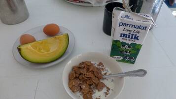 A breakfast setup with a bowl of cereal, a plate with an avocado slice and eggs, a carton of milk, and a metal container.
