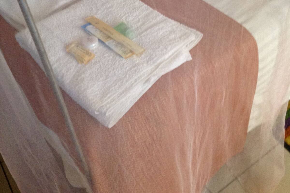 Ein Bett mit einer rosa Bettdecke und einem weißen Kopfkissen. Auf dem Bett liegt ein weißes Handtuch zusammen mit einer kleinen Flasche und einem Taschentuchpaket.
