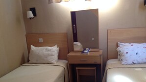 Habitació d'hotel petita amb dos llits individuals, una tauleta de nit de fusta amb mirall, una taula petita i una làmpada de paret.