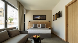 A hotel room with a bed, a sofa, a small table with a fruit bowl, and a window with curtains. - Fos DownTown Suites (Athens)