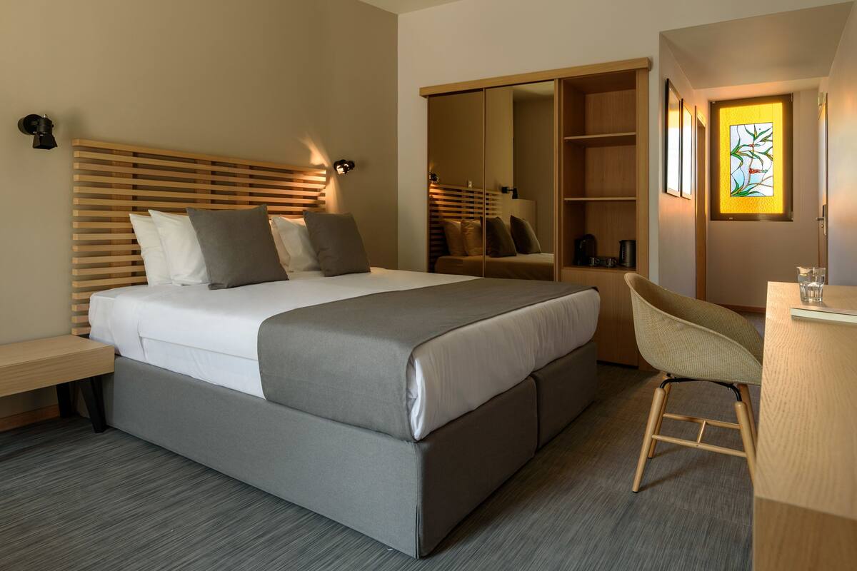 A modern hotel room with a large bed, a desk, a chair, and a wardrobe.