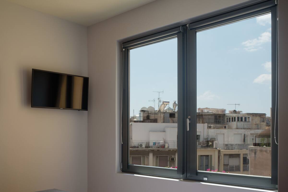 A bedroom with a bed, a TV mounted on the wall, and a large window with a city view.