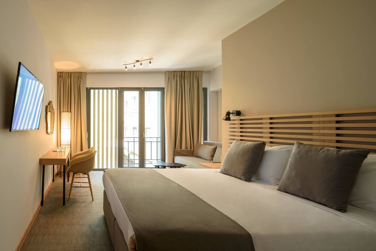 A hotel room with a large bed, a desk with a chair, a television, and a balcony with curtains.