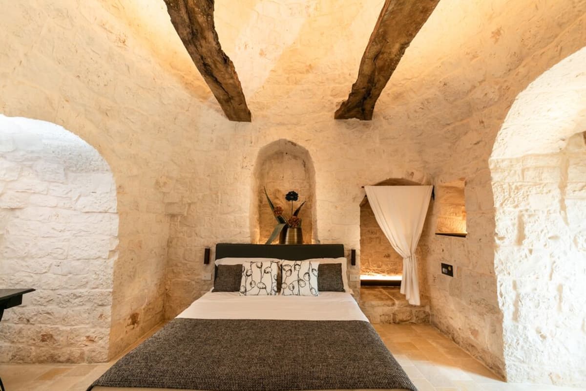 Una habitación rústica de piedra con una cama, una mesita pequeña y un jarrón decorativo.