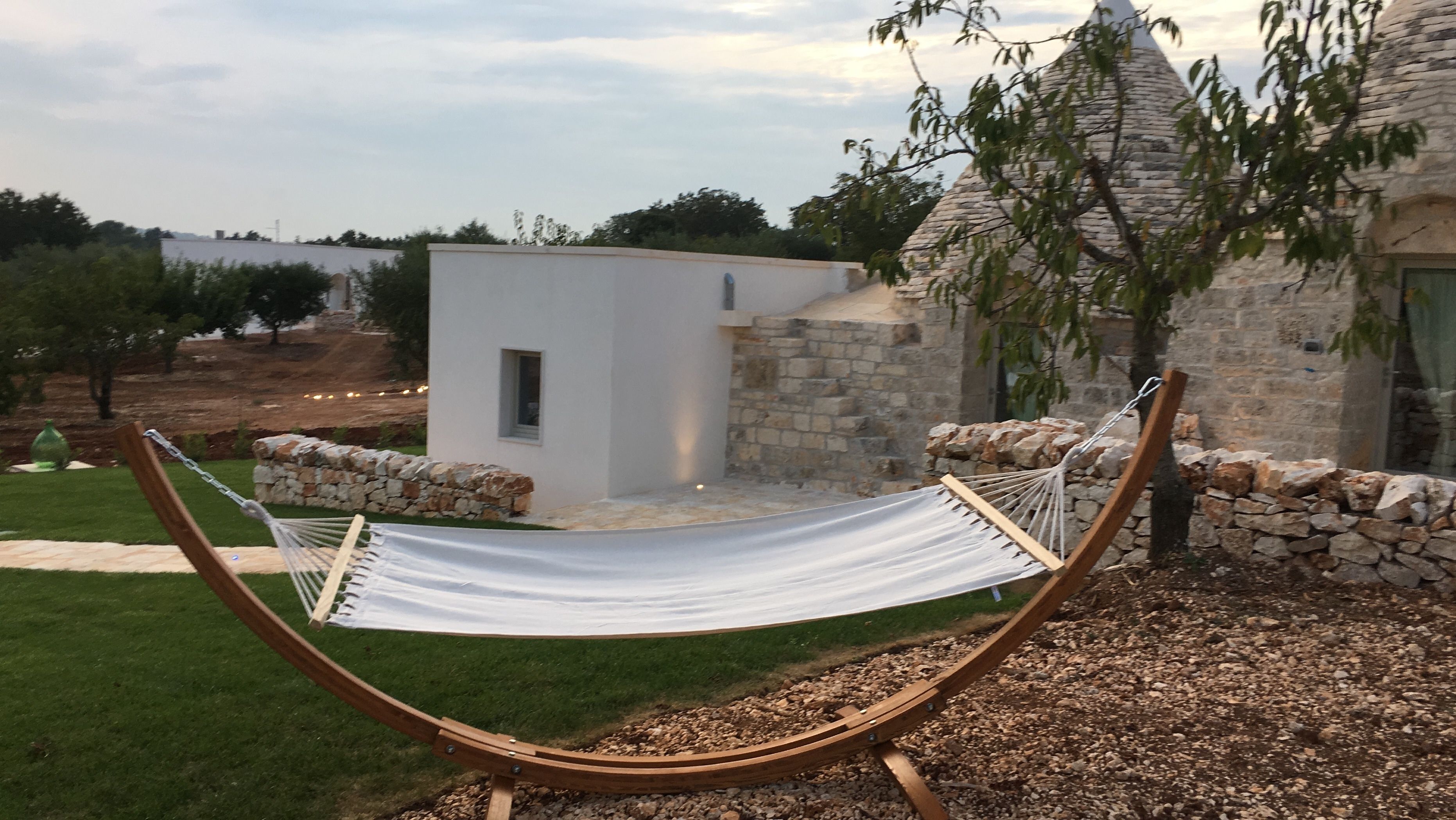 A hammock in a garden area with a stone wall and a small white building in the background.