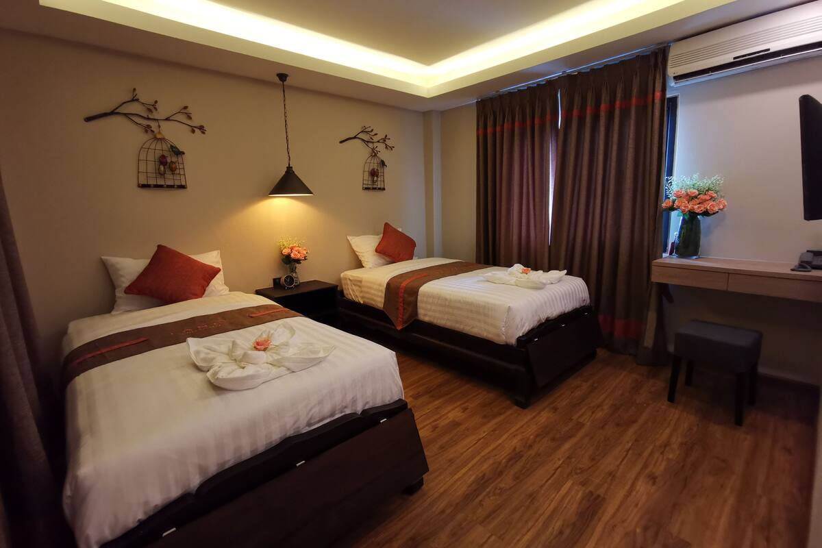 A hotel room with two beds, a desk, and a chair. There are decorative wall pieces and a vase with flowers on the desk.