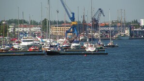 A marina with multiple boats docked, cranes, and industrial buildings in the background.