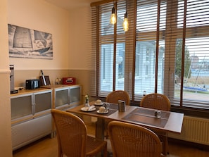 A dining area with a table set for four, wicker chairs, a radiator, a window with blinds, and a wall-mounted picture. - Hotel Pension Zum Alten Fährhaus (Rostock)