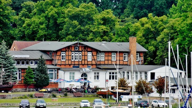 A traditional half-timbered building with a sign for a restaurant, boats docked in the foreground, and a forested hill in the background.