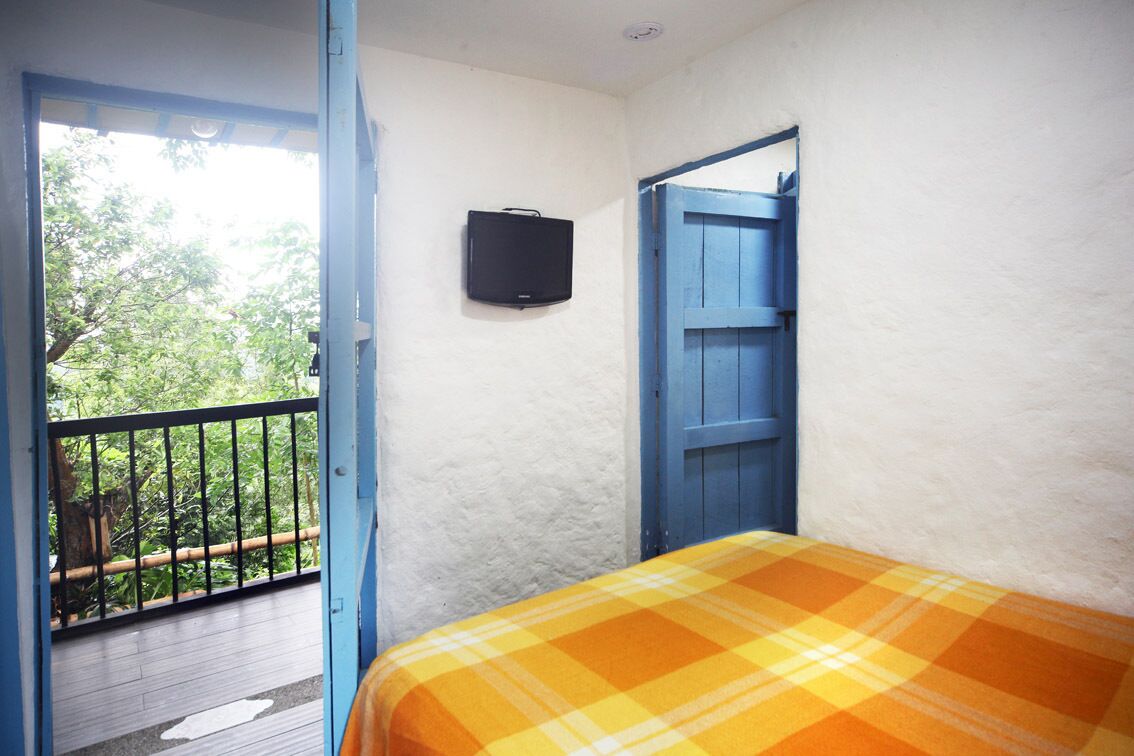 Una habitación con una cama, un televisor, una puerta azul y un balcón con barandilla.