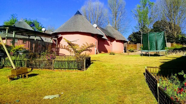 Une maison traditionnelle à toit de chaume, peinte en rose, entourée d’un jardin bien entretenu où poussent différentes espèces de plantes et délimitée par une clôture en bois.