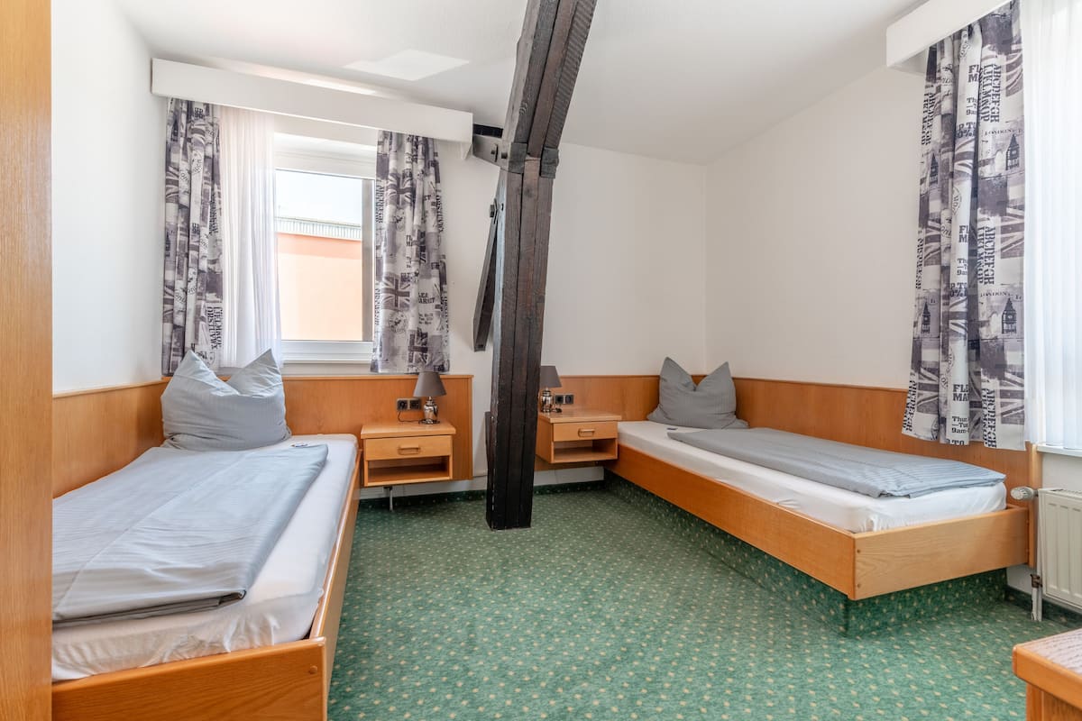 Habitación de hotel con dos camas individuales, un armario de madera, una mesita de noche con lámpara, una ventana con cortinas estampadas y un radiador.