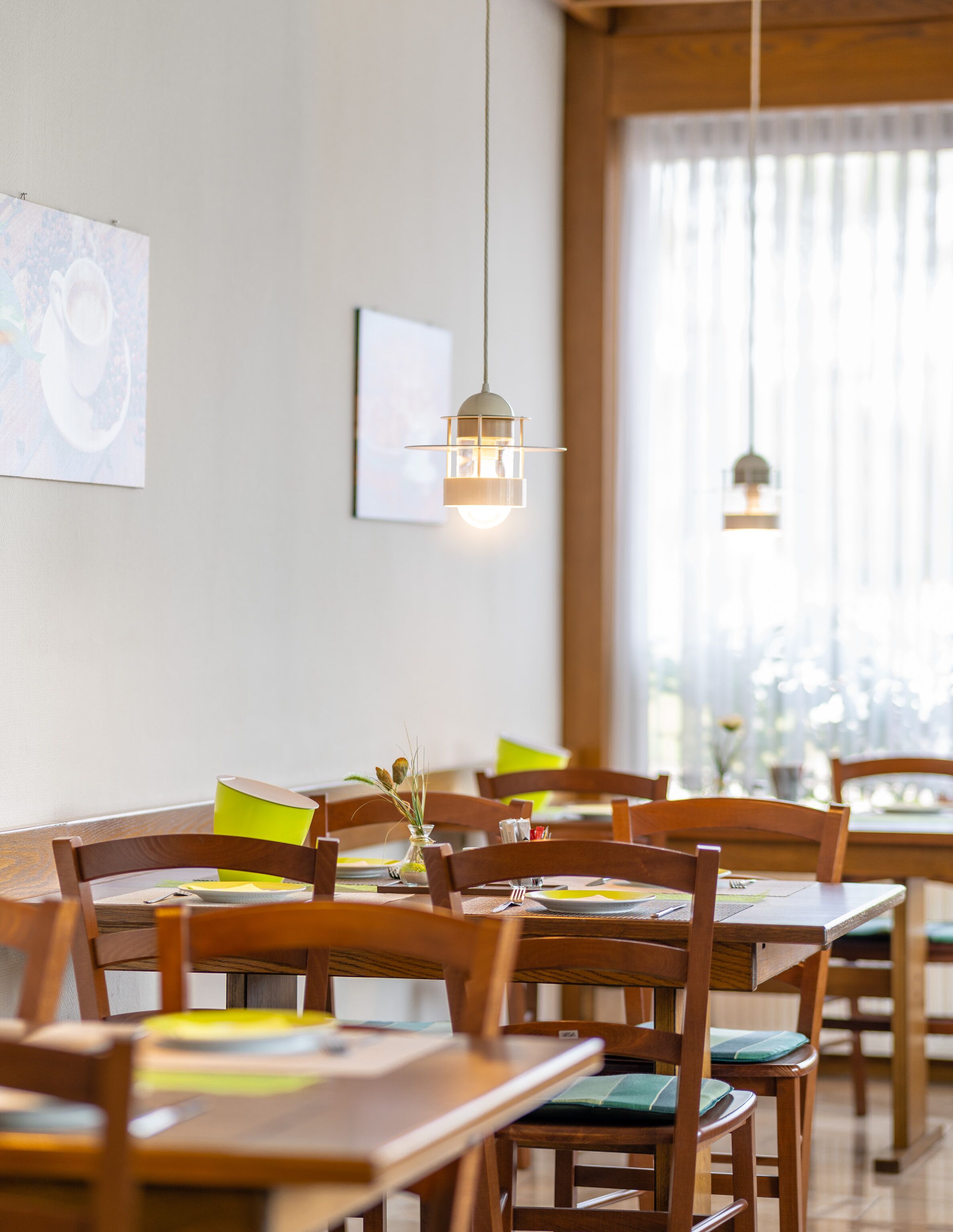 A dining area with wooden tables and chairs, a hanging light fixture, and a window with sheer curtains.