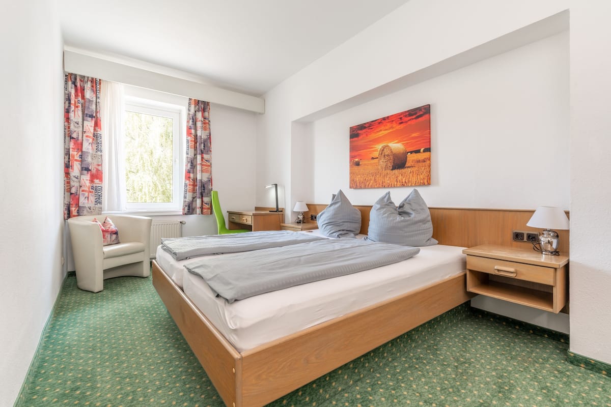 Una habitación de hotel con una cama, una silla, un escritorio, una lámpara y un cuadro en la pared.