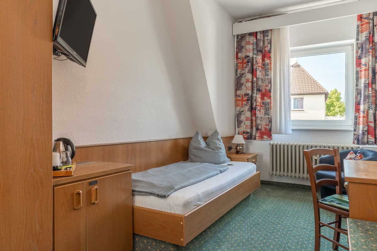 Una habitación pequeña y luminosa con una cama, un escritorio, una silla, un televisor montado en la pared y un ventanal con cortinas.