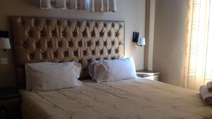 A hotel room with a large bed, two nightstands, a wall-mounted lamp, and a window with curtains. - Little Lodge (Choma)