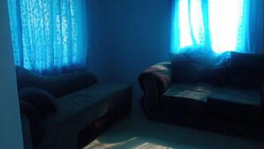 A room with two sofas and a bed, a window with blue curtains, and a wall-mounted light fixture. - Little Lodge (Choma)