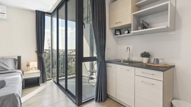Un appartement moderne doté d’un balcon, d’une cuisine avec des meubles blancs et d’un lit avec une tête de lit grise.