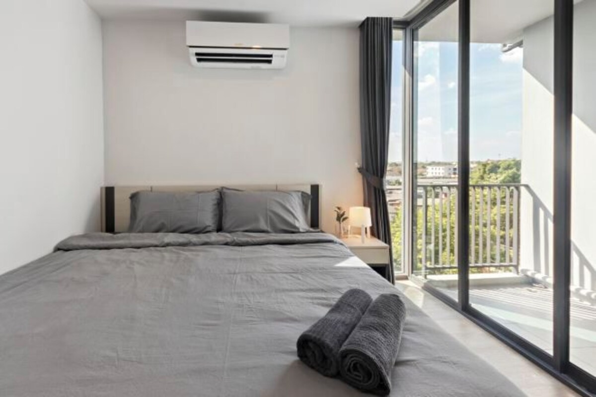 A modern hotel room with a large bed, grey bedding, a window with a view of the city, and an air conditioning unit.