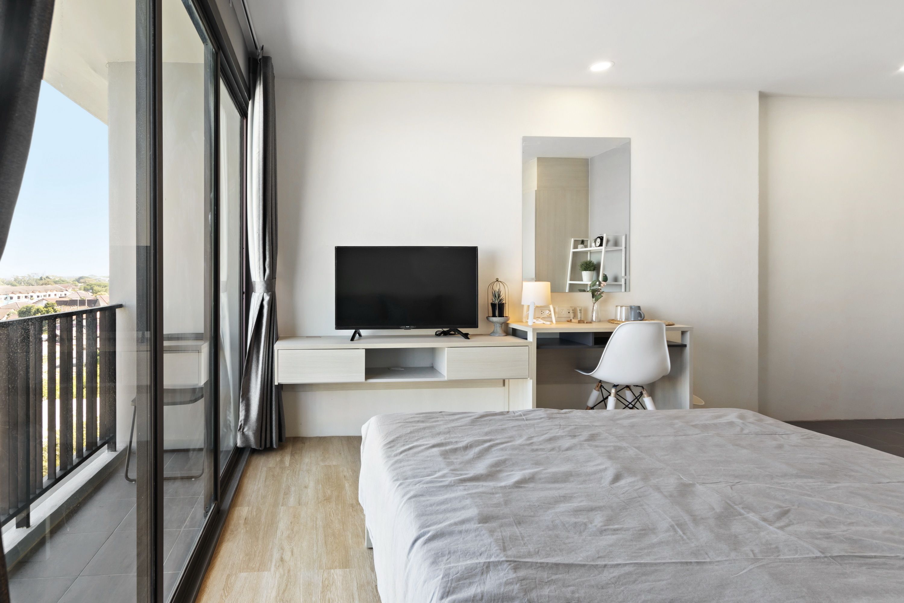 A modern hotel room with a large bed, a desk with a flat-screen TV, a chair, a small table with a lamp, and a large window with a balcony.