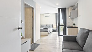 Une pièce moderne et bien éclairée, avec un canapé gris, un lit avec une literie grise, un petit coin cuisine équipé d’un évier et d’un plan de travail, et une grande fenêtre avec des rideaux.