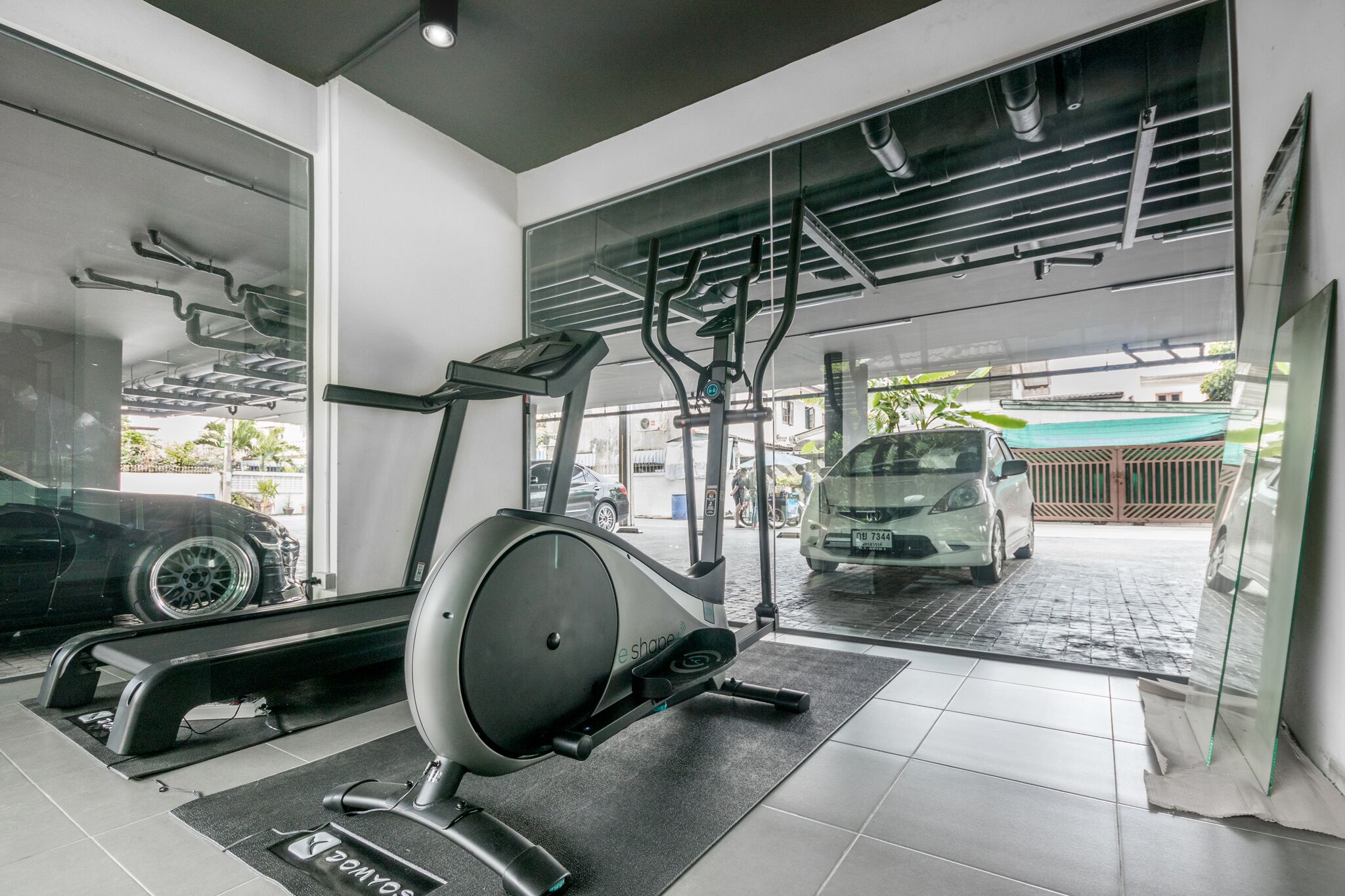 Un gimnasio con equipo de ejercicio, un coche al fondo y vistas al exterior a través de amplias ventanas.
