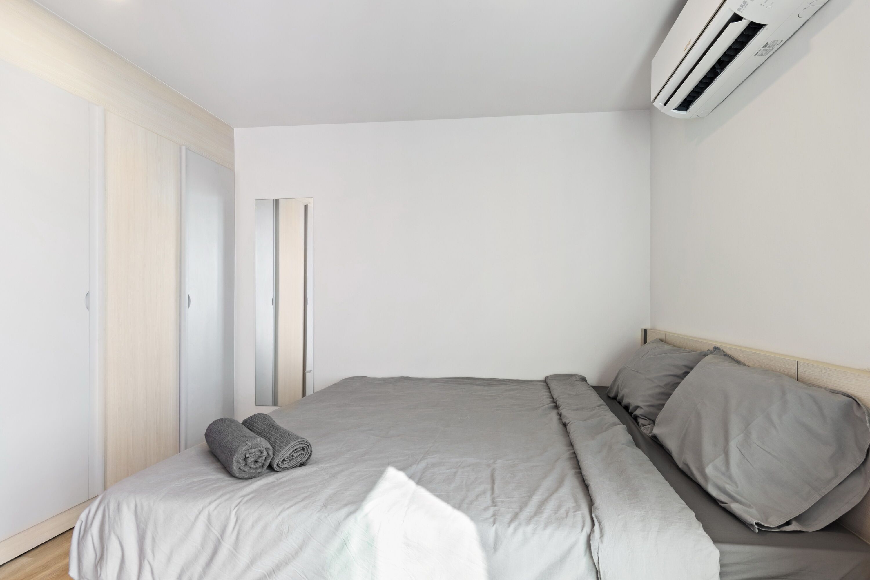 Un dormitori minimalista amb llit, roba de llit gris, aire condicionat fixat a la paret i un armari de fusta.