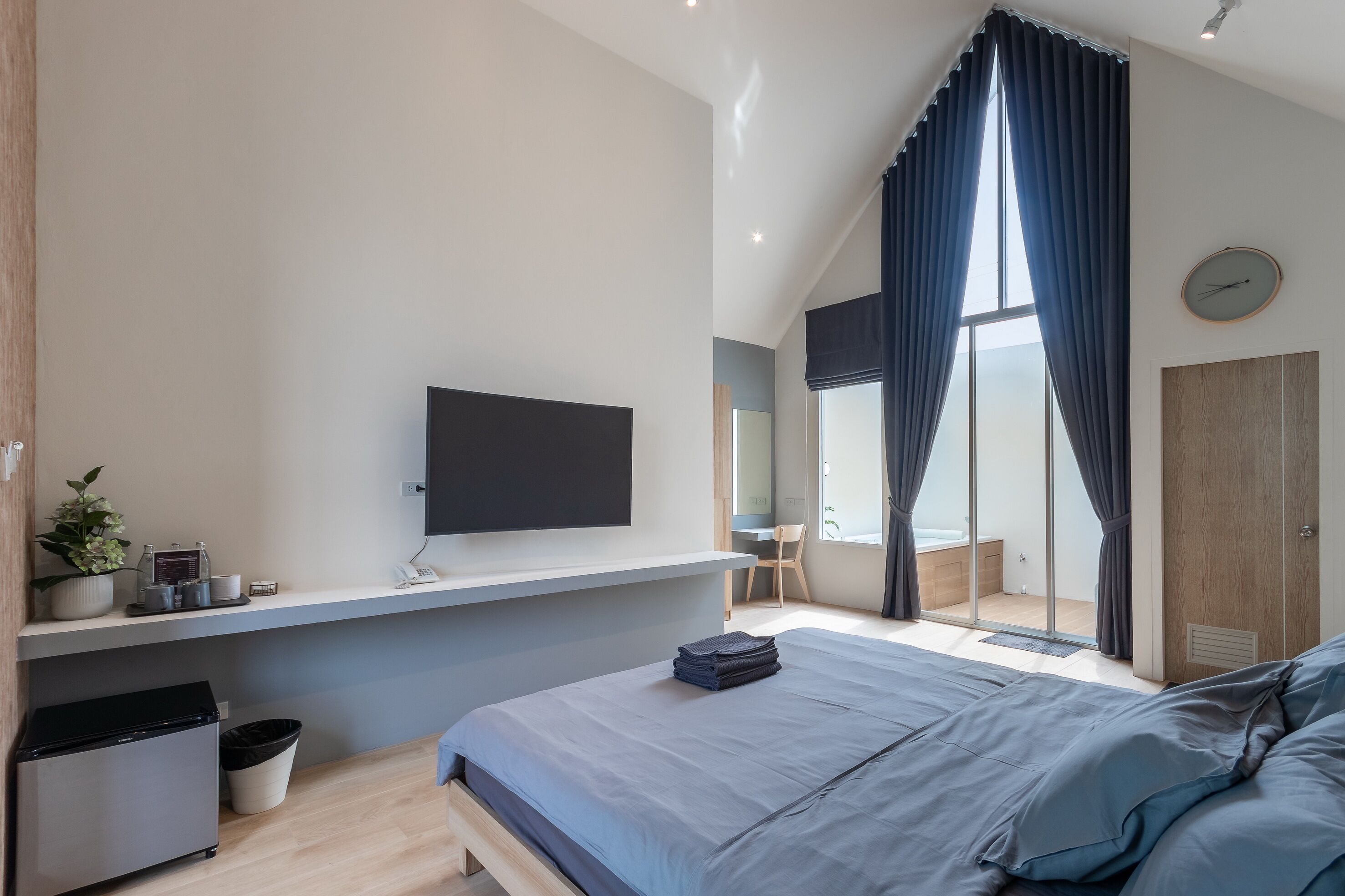 A modern bedroom with a bed, a TV mounted on the wall, a desk, and a window with curtains.