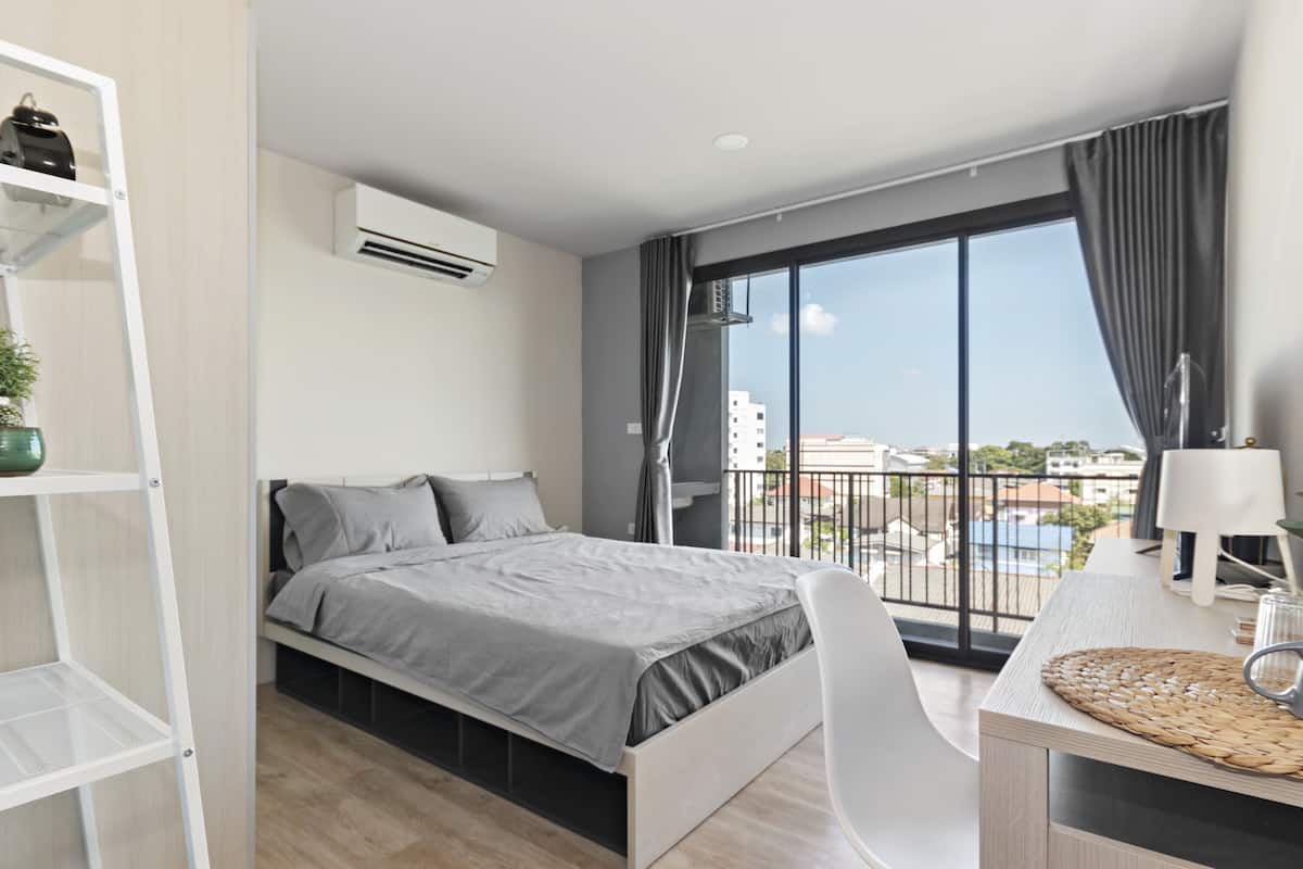 A modern bedroom with a bed, a desk, and a large window offering a city view.