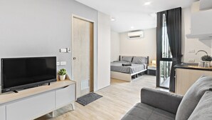 Une pièce moderne et bien éclairée, avec un téléviseur à écran plat posé sur un meuble télé en bois, un canapé gris, un lit avec une literie grise et un petit coin cuisine équipé d’un évier et de placards.