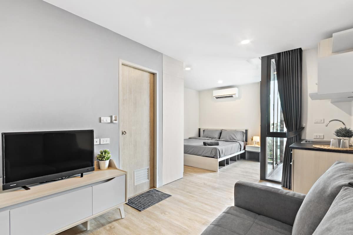 A modern, well-lit room with a flat-screen TV on a wooden TV stand, a grey sofa, a bed with grey bedding, and a small kitchen area with a sink and cabinets.
