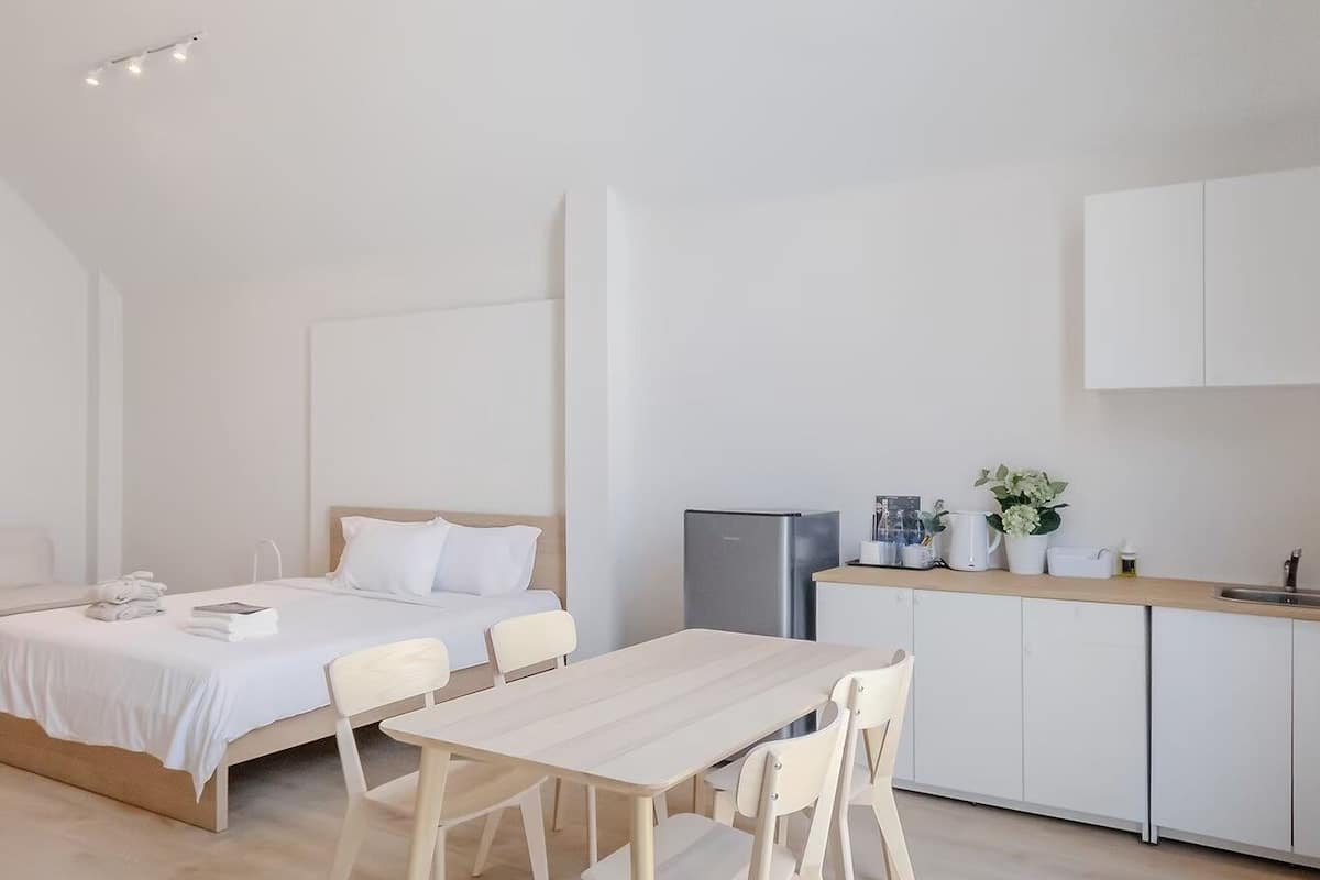 A modern, minimalist bedroom with a large bed, a wooden dining table, and a kitchen area.