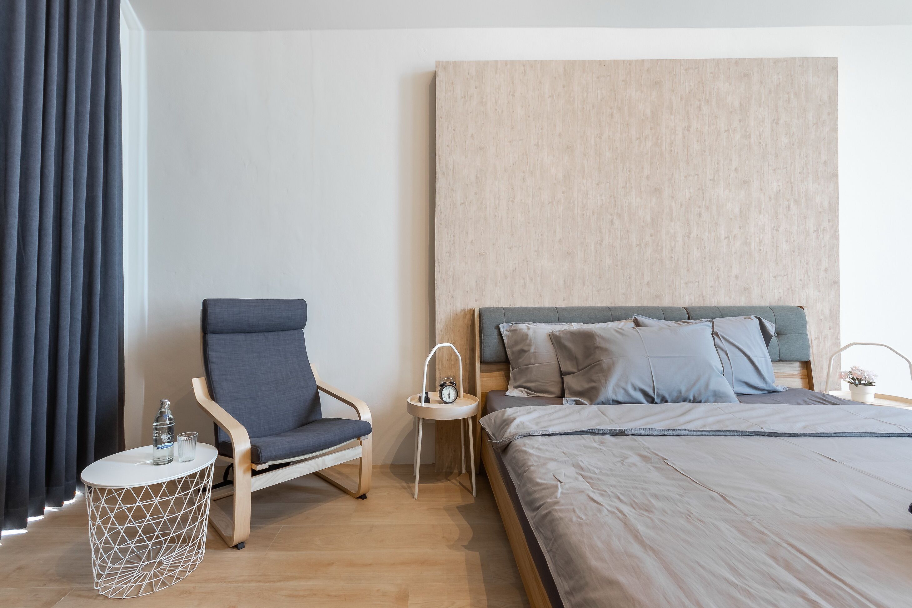A modern bedroom with a bed, a chair, a side table with a clock, and a curtain.