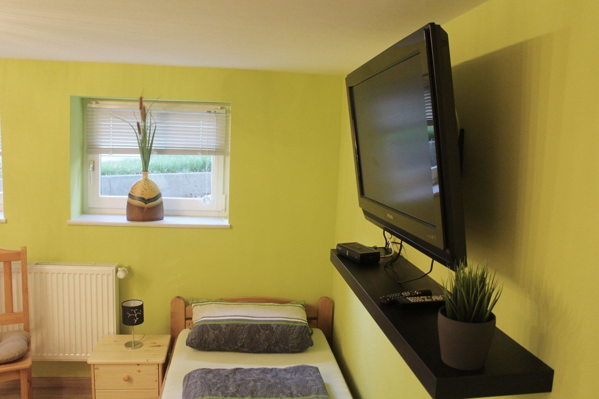 A room with a bed, a television on a stand, a window with blinds, a radiator, a nightstand, and a potted plant.