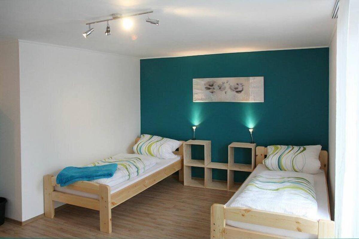 A room with two wooden beds, a teal accent wall, and a ceiling-mounted light fixture.