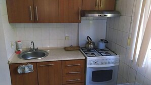 Een kleine keuken met houten kastjes, een roestvrijstalen spoelbak, een gasfornuis en een witte tegelachterwand.