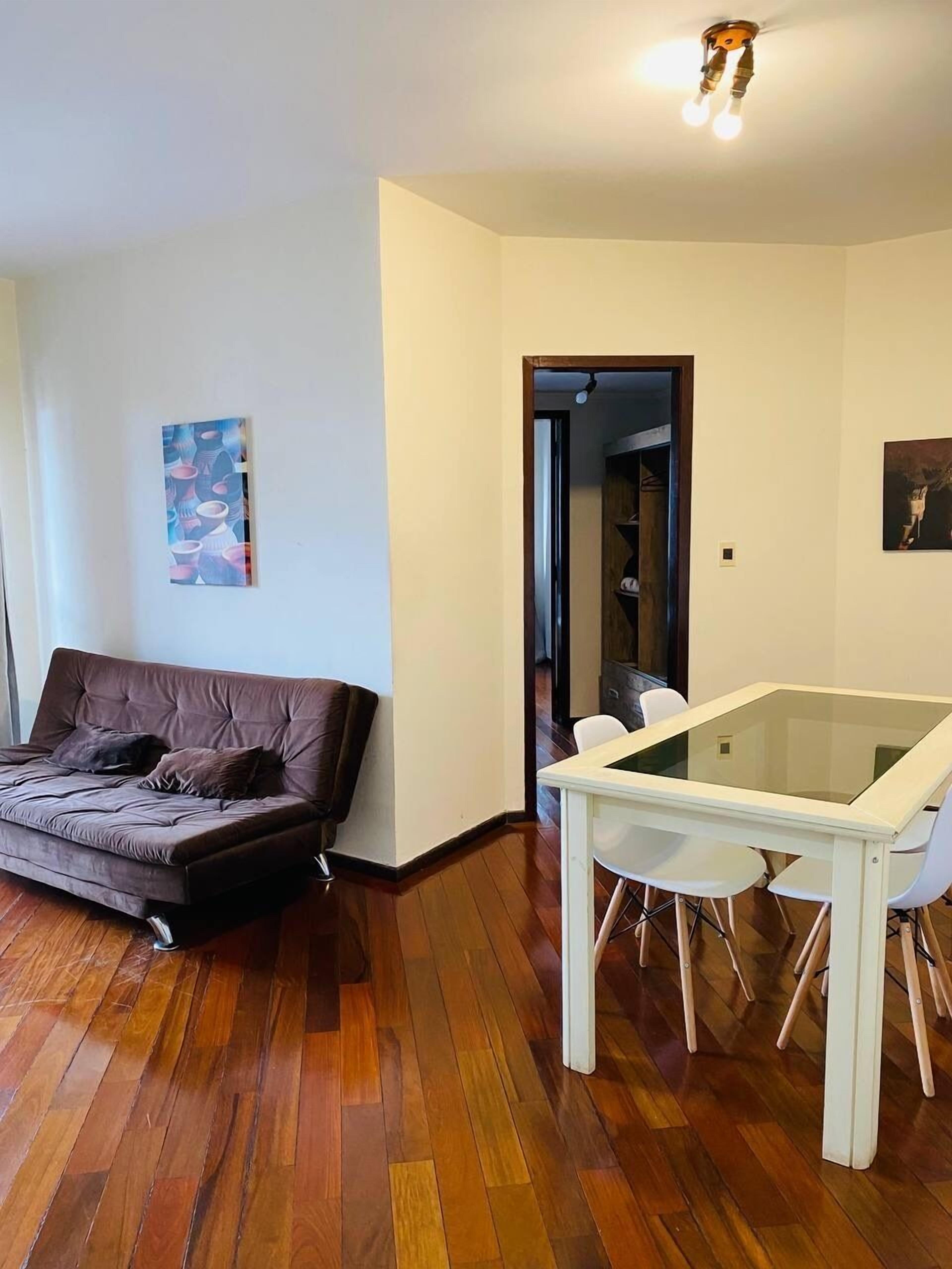 A modern living room with a brown sofa, a glass-top dining table with white chairs, wooden flooring, and a painting on the wall.