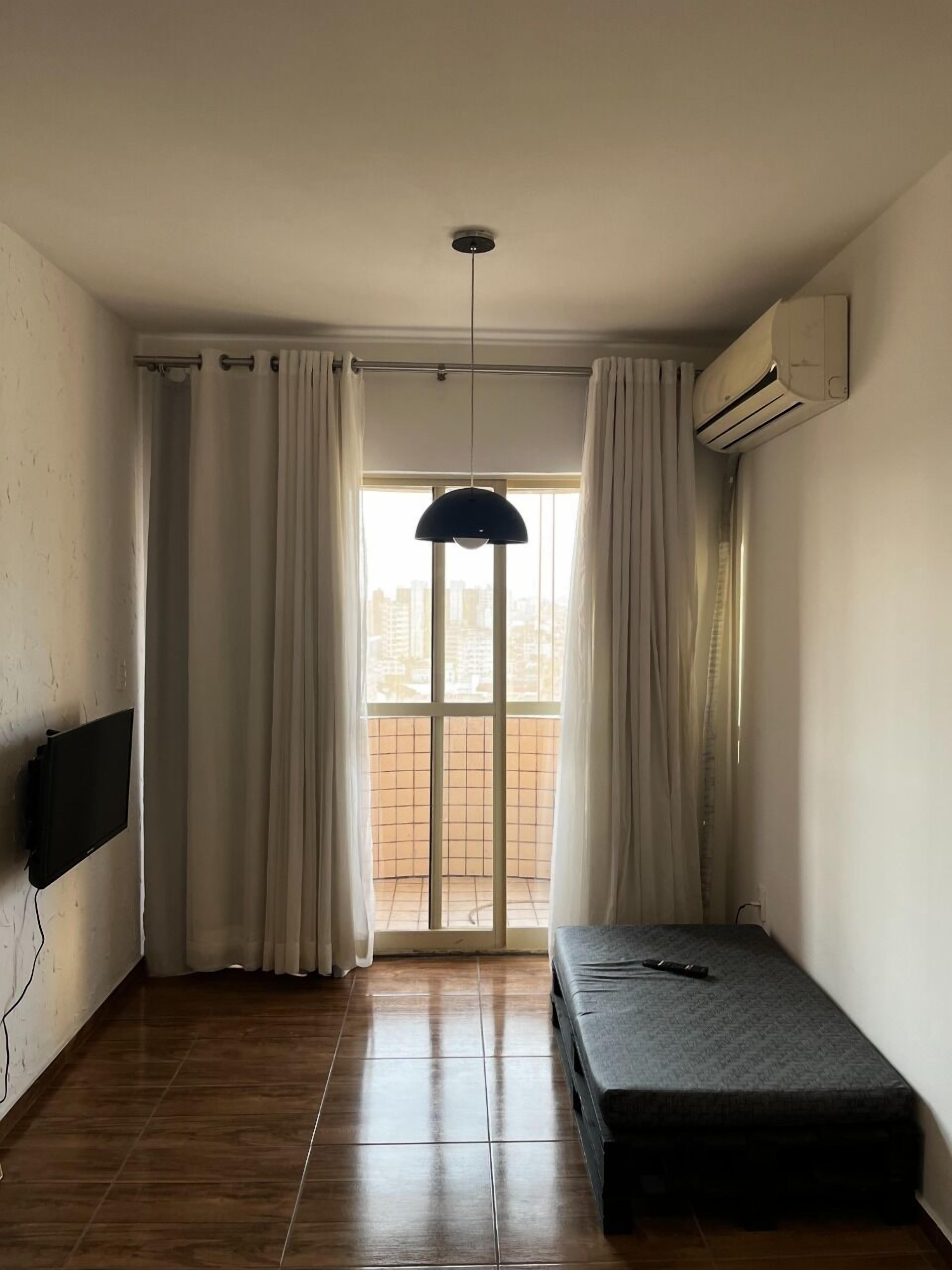 A small, sparsely furnished room with a single bed, a mounted TV, and a large window with curtains.