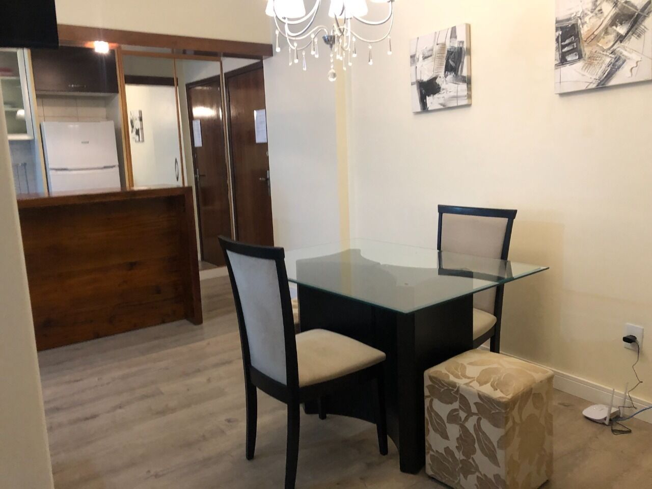 A dining area with a glass table, two chairs, a bench, a chandelier, and artwork on the wall.