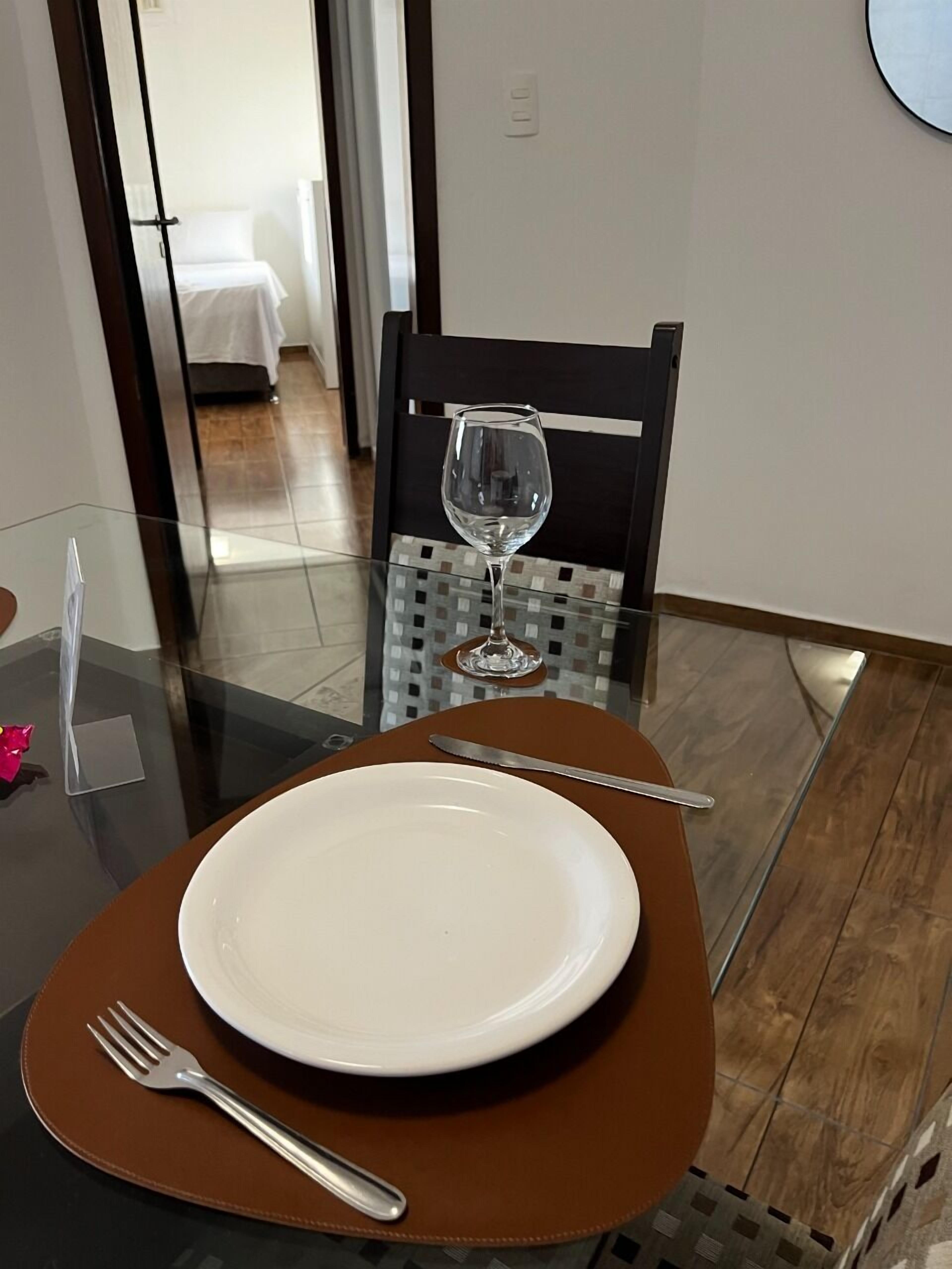 Una mesa de comedor con un plato, un tenedor y una copa de vino, ubicada en una habitación con una silla y vista a una cama al fondo.