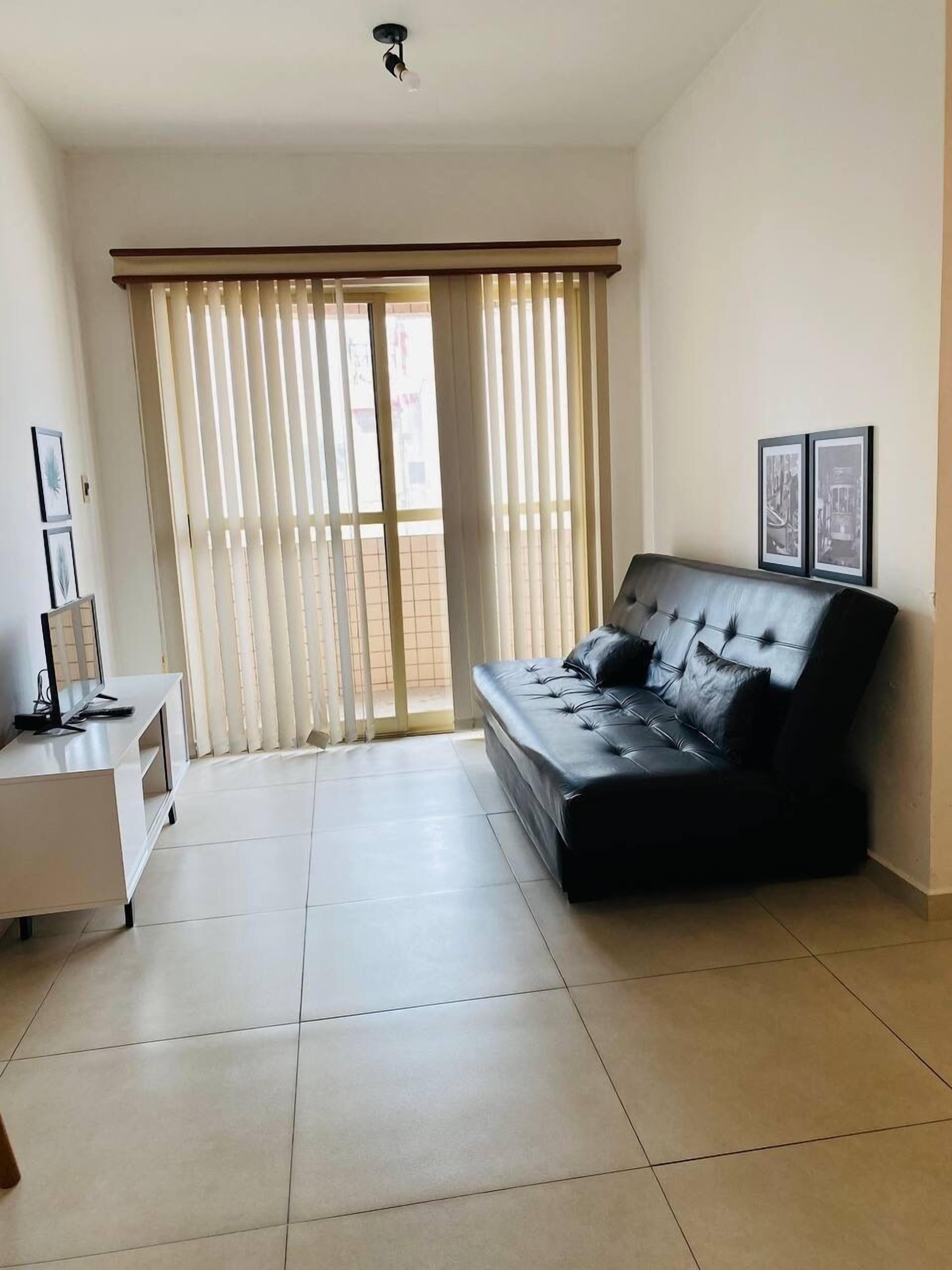 Una sala de estar moderna con un sofá de cuero negro, un mueble blanco para entretenimiento y un ventanal con persianas verticales.