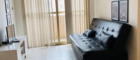 Una sala de estar moderna con un sofá de cuero negro, un mueble blanco para entretenimiento y un ventanal con persianas verticales.