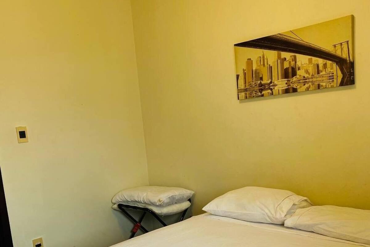 Hotellihuone, jossa on sänky, tyynyjä, taiteltu pyyhe ja kaupunkimaisemaa esittävä taulu seinällä.