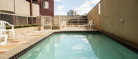 A swimming pool with white chairs and a fence, surrounded by buildings.