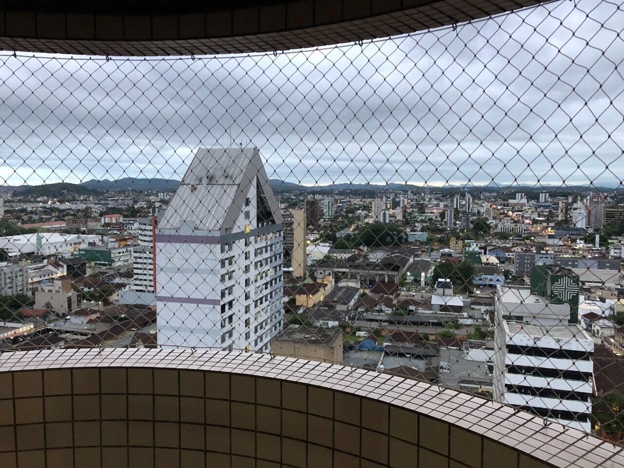 A cityscape viewed through a chain-link fence, featuring a prominent white building with a triangular roof and a smaller adjacent structure.
