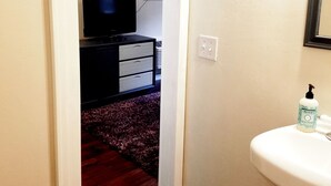 Un couloir mène à une chambre équipée d’une télévision, d’une commode et d’une salle de bain avec un lavabo et du savon liquide.