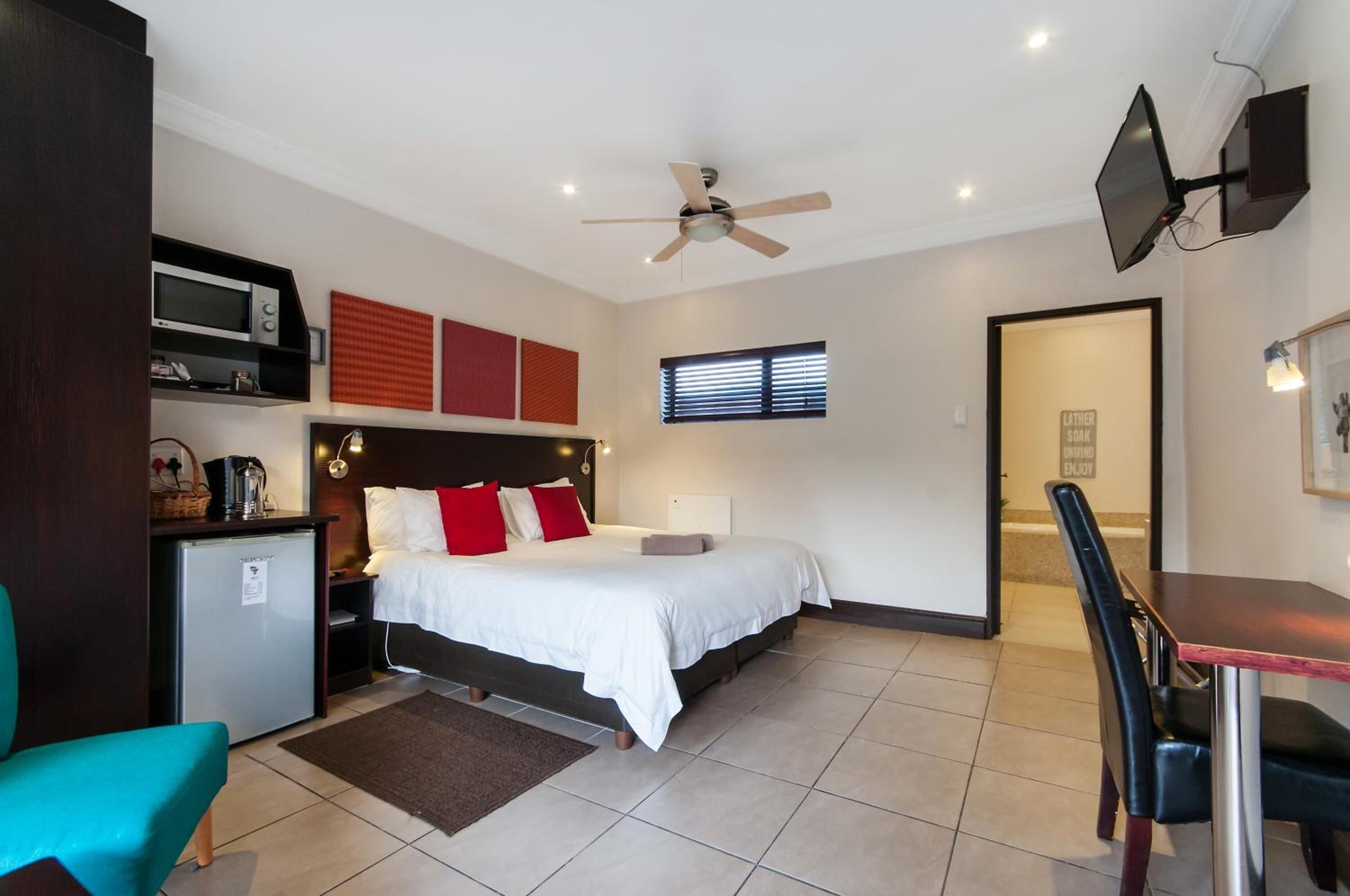 Habitación de hotel compacta con cama, un escritorio pequeño con silla, ventilador de techo, televisor montado en la pared y ventana con persianas.