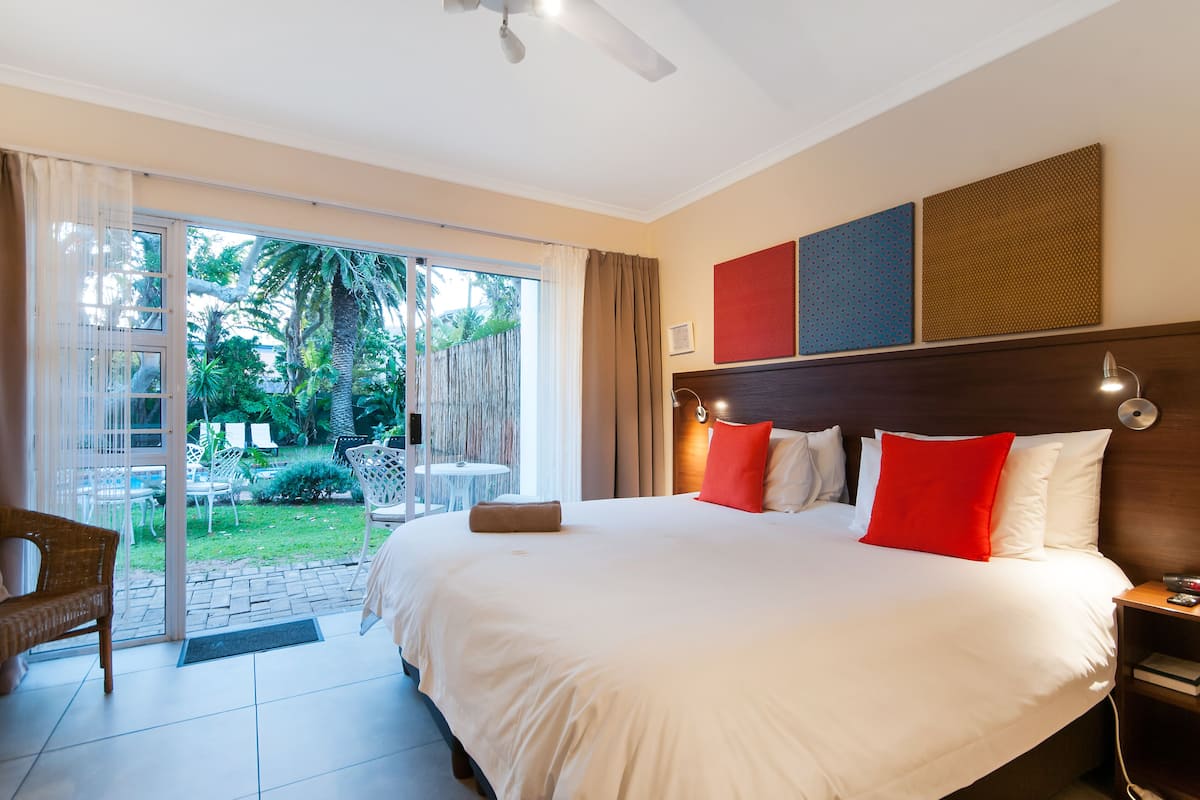Habitación de hotel con una cama grande, cabecera de madera, mesita de noche y vista a un jardín a través de una puerta corrediza de vidrio.